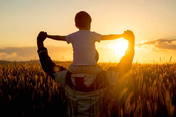 Père portant son enfant sur les épaules dans un champ face au soleil